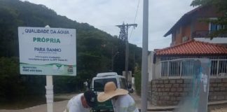 dois garis retiram saco de lixo cheio de três lixeiras de concreto na areia da praia com placa de qualidade própria da água ao lado