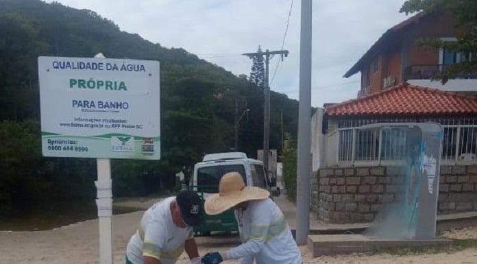 dois garis retiram saco de lixo cheio de três lixeiras de concreto na areia da praia com placa de qualidade própria da água ao lado