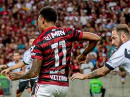 jogadores de flamengo e vasco se movimentando em campo com torcida ao fundo