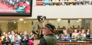 mafalda visto de costas empunha ao alto uma câmera fotográfica no plenário da alesc lotado de pessoas aplaudindo