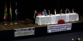 auditório visto lotado com mesa bancada no palco com pessoas sentadas, faixas à frente e bandeiras ao lado