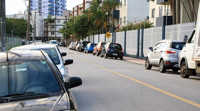 carros estacionados nos dois lados de uma rua asfaltada com prédios em volta