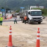 caminhões e operários sobre a pista onde há cones e montes de areia