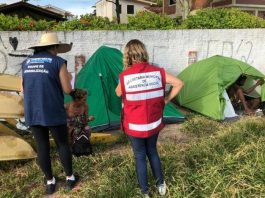 duas mulheres com uniformes da prefeitura vistas de costas conversando com moradora de rua ao lado de duas barracas em área de grama alta ao lado de um muro