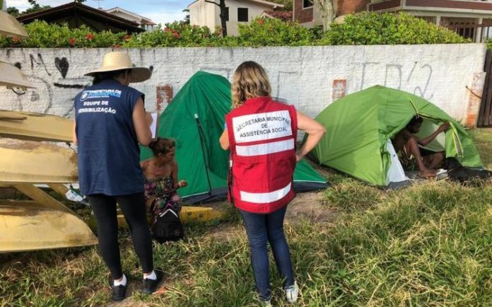 duas mulheres com uniformes da prefeitura vistas de costas conversando com moradora de rua ao lado de duas barracas em área de grama alta ao lado de um muro