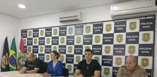 bancada com quatro pessoas: a delegada, um delegado, um policial municipal e um policial militar, com painel de logos da polícia civil e bandeiras ao fundo