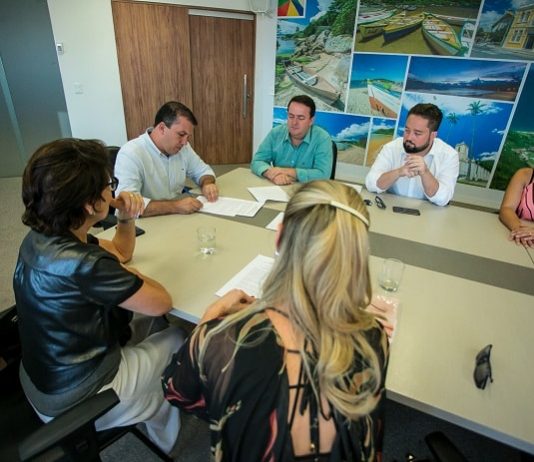 na ponta de uma mesa o prefeito camilo martins assina documento com outros homens e mulheres sentados em volta olhando