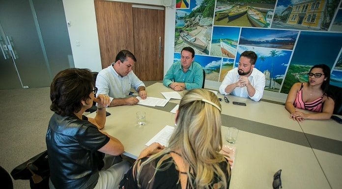 na ponta de uma mesa o prefeito camilo martins assina documento com outros homens e mulheres sentados em volta olhando