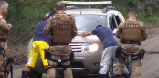 PM prende em Laguna suspeito de estupro em Santo Amaro da Imperatriz dois homens apoiados com as mãos em viatura e policiais em volta