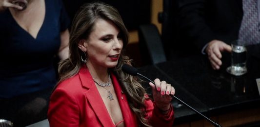 deputada paulinha discursando ao microfone com a roupa decotada na cerimômia de posse