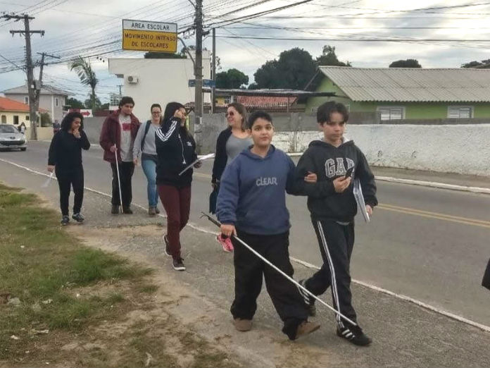 garoto cego com bengala acompanhado pelo braço por outro garoto andando com mais pessoas em um acostamento
