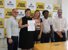 as cinco pessoas em pé posam para foto; adeliana mostra um papel nas mãos; ao fundo banner do projeto mãos à obra