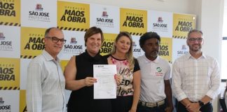 as cinco pessoas em pé posam para foto; adeliana mostra um papel nas mãos; ao fundo banner do projeto mãos à obra