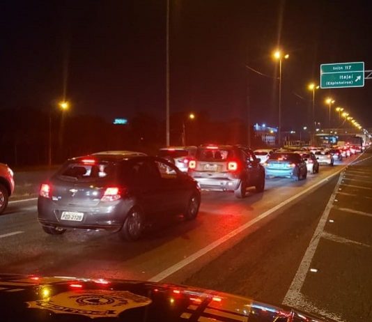 veículos parados em fila nas duas pistas da br 101 com placa para itajaí e viatura no acostamento