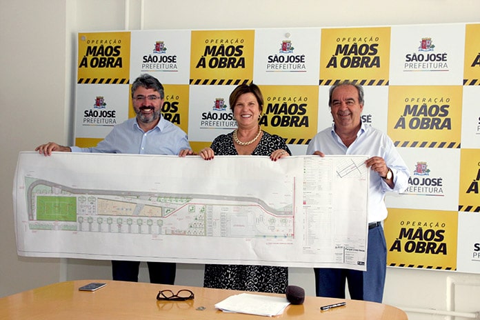 sanderson adelina e neri posam em pé para foto segurando uma grande tira de papel mostrando o projeto técnico da praça com painel ao fundos com logos da prefeitura e inscrições "mãos à obra"