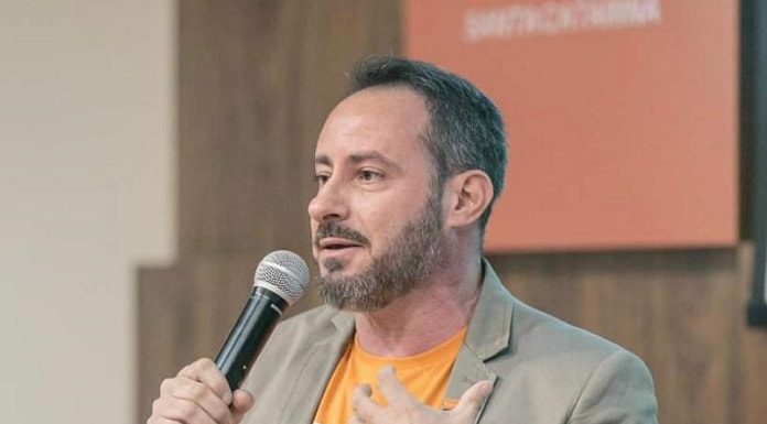 victor souza discursa em pé com um microfone na mão, usando jaqueta e por baixo camiseta laranja do partido