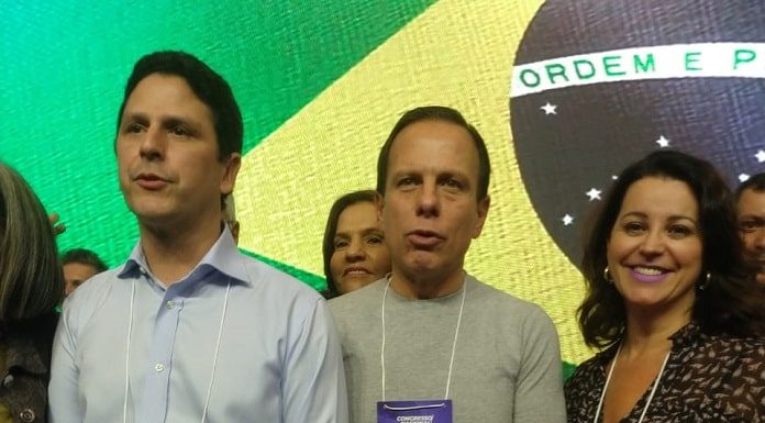 três pessoas lado a lado com a bandeira do brasil em telão ao fundo