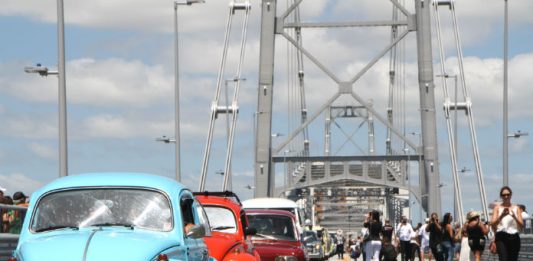 fuscas e outros carros antigos passando sobre a ponte Hercílio Luz com pessoas em volta