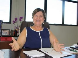 adeliana olha para a câmara sorridente e de braços abertos sentada em mesa de seu gabinete