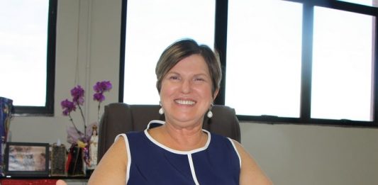 Prefeita Adeliana encerra 2019 comemorando investimentos adeliana olha para a câmara sorridente e de braços abertos sentada em mesa de seu gabinete