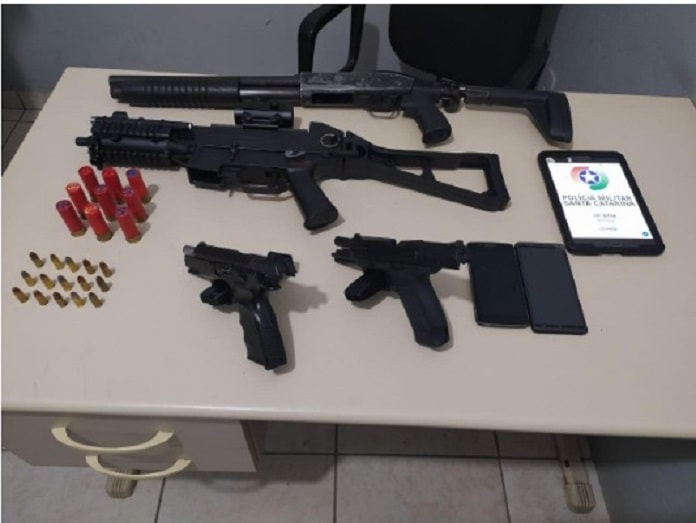 armas dispostas sobre uma mesa com as munições, celulares e um tablet com logo da pm ao lado