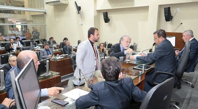 vereadores conversam em volta da mesa diretora da câmara, alguns em pé na frente e outros sentados; plenário ao fundo