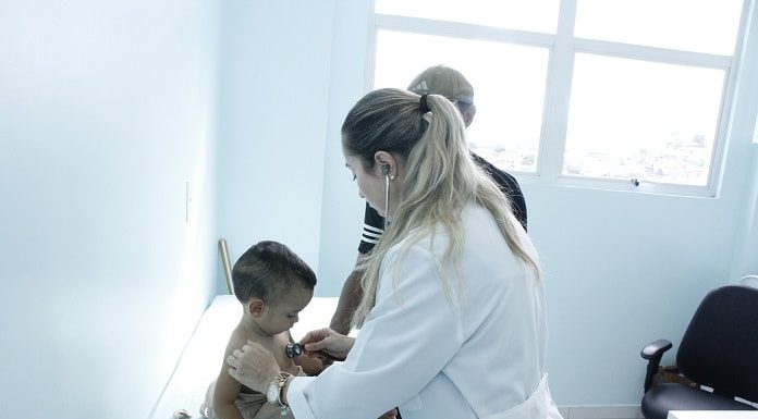 médica ausculta peito de criança sentada em maca com outra pessoas atrás