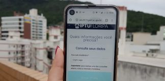 mulher segura celular com site do iptu aberto sobre um prédio com outros prédios ao fundo