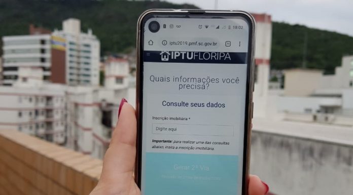 mulher segura celular com site do iptu aberto sobre um prédio com outros prédios ao fundo