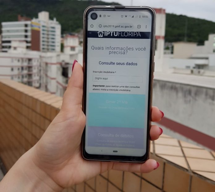 mulher segura celular com site do iptu aberto sobre um prédio com outros prédios ao fundo