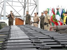 PM recebe 100 fuzis usados do exército brasileiro fuzis enfileirados em primeiro plano com militares do exército e da polícia ao fundo, bandeiras e uma estrutura da ponte hercílio luz