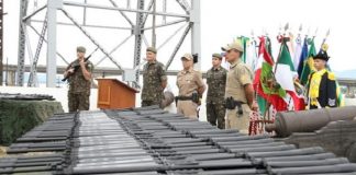 fuzis enfileirados em primeiro plano com militares do exército e da polícia ao fundo, bandeiras e uma estrutura da ponte hercílio luz