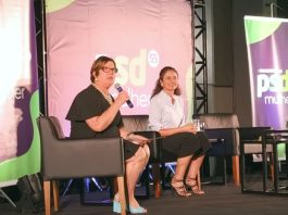 PSD reúne 250 mulheres para debater a participação feminina na política adeliana e marlene sentada em poltronas em um palco. adeliana segura um microfone; há um banner do partido ao fundo