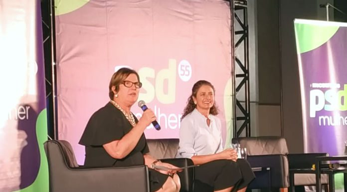 adeliana e marlene sentada em poltronas em um palco. adeliana segura um microfone; há um banner do partido ao fundo