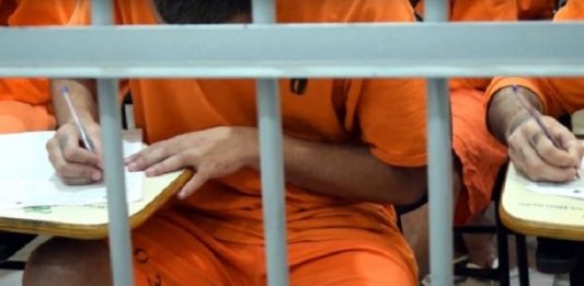 preso com uniforme laranja fazendo prova atrás de grades