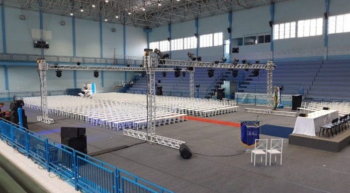 centenas de cadeiras brancas no centro de um ginásio com estruturas de iluminação ao redor e pequeno palco; não há ninguém na foto