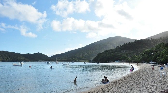 praia de mar calmo, cinco pessoas na praia, alguns barcos e morros de mata preservada ao redor