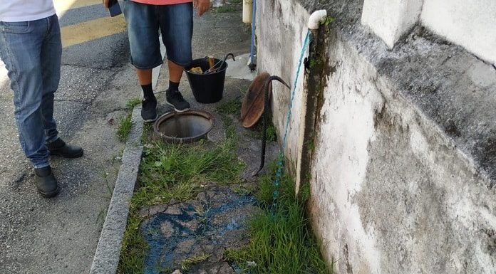 líquido azul escorrendo pela calçada a partir de cano no muro e um bueiro aberto ao lado, com dois fiscais em volta