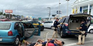 quatro homens (três sem camisa) algemados deitados com o rosto contra o asfalto ao lado do carro na marginal com viaturas em volta