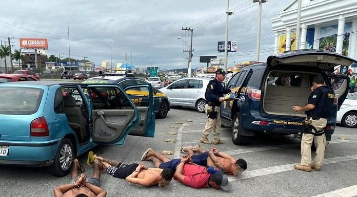 quatro homens (três sem camisa) algemados deitados com o rosto contra o asfalto ao lado do carro na marginal com viaturas em volta