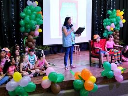 Projeto Troca Troca Literário, do Melão, celebra 24 anos professora lê em pé no palco com diversas crianças sentadas à sua volta e fantasiadas