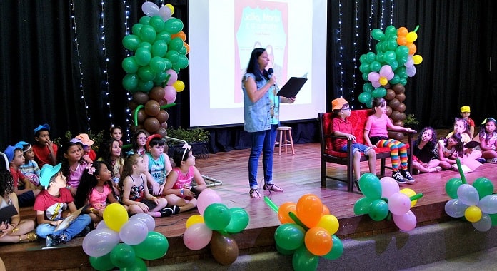 professora lê em pé no palco com diversas crianças sentadas à sua volta e fantasiadas