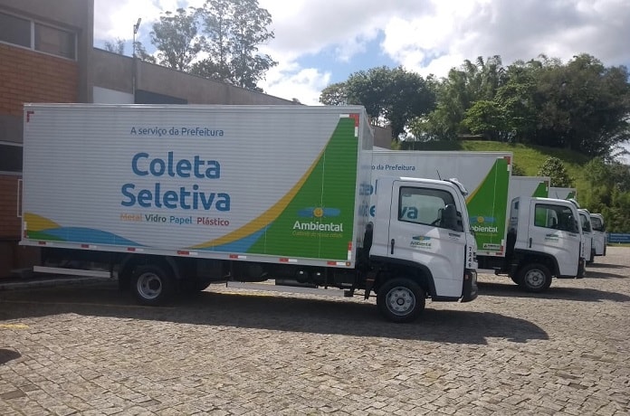 quatro caminhões de coleta de lixo seletiva com logo da ambiental estacionados lado a lado de ré para um edifício