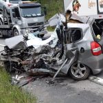 honda fit completamente destruído em acostamento da rodovia e caminhões passando ao lado