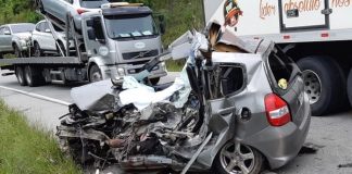 honda fit completamente destruído em acostamento da rodovia e caminhões passando ao lado