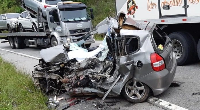 honda fit completamente destruído em acostamento da rodovia e caminhões passando ao lado