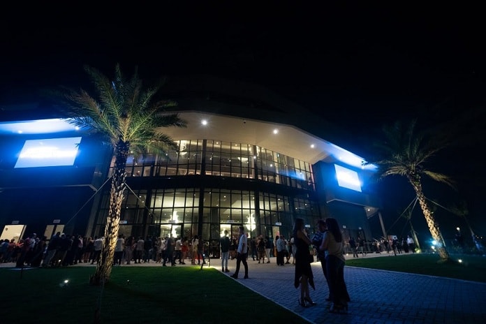 fachada iluminada da arena petry com dezenas de pessoas na frente em foto noturna
