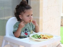 menina comendo em cadeirote infantil com prato de legumes a sua frente