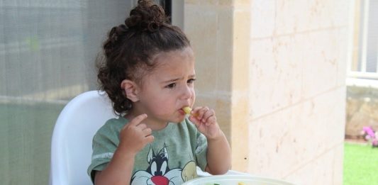Como introduzir uma alimentação balanceada na rotina das crianças menina comendo em cadeirote infantil com prato de legumes a sua frente