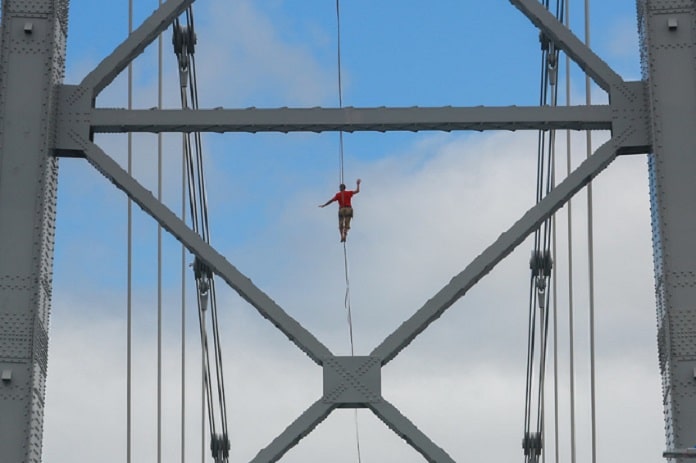 homem em pé sobre a fita no alto visto através das estruturas de uma torre da ponte
